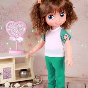 以下が含まれることがあります： 茶色の巻き毛と青い目の人形。白いTシャツに緑色の袖と緑色のズボンを着用しています。人形にはそばかすがあり、小さなピンクの花のヘアクリップを付けています。背景には、クリスタルが付いたピンクのハート型の装飾があります。