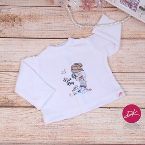 May include: White long-sleeve shirt with a graphic of a girl wearing a brown hat and holding a camera. The text "Nice Day" is printed on the shirt.