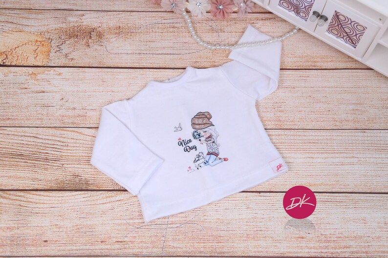 May include: White long-sleeve shirt with a graphic of a girl wearing a brown hat and holding a camera. The text "Nice Day" is printed on the shirt.
