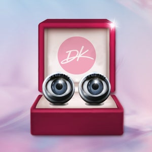 May include: A red velvet box with a pink circle containing the letters "DK" in white. The box contains two silver-colored eyeball cufflinks with black eyelashes.