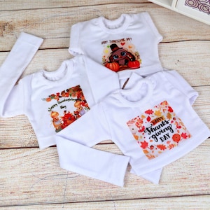 May include: Three white baby shirts with Thanksgiving-themed graphics. One shirt has a long sleeve. The graphics include fall leaves, pumpkins, and the phrases "Happy Thanksgiving Day" and "Happy Thanks Giving Day".