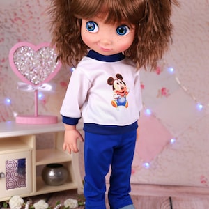 May include: A doll dressed in a white top with a Mickey Mouse print, blue trousers, and denim trainers. The doll has brown curly hair, blue eyes, and blue flower hair clips. It stands before a pink heart-shaped decoration.