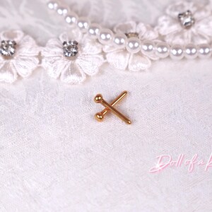 May include: A small, gold-colored metal pin with two crossed needles, each with a ball on the end. The pin is on a white, textured fabric, with a pearl necklace and lace trim with rhinestones in the background.