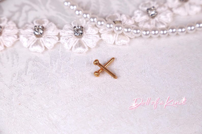 May include: A small, gold-colored metal pin with two crossed needles, each with a ball on the end. The pin is on a white, textured fabric, with a pearl necklace and lace trim with rhinestones in the background.