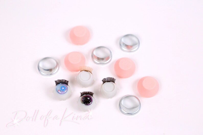 May include: Assortment of doll eye supplies including clear plastic eye sockets, pink silicone eye sockets, and two sets of doll eyes with lashes.