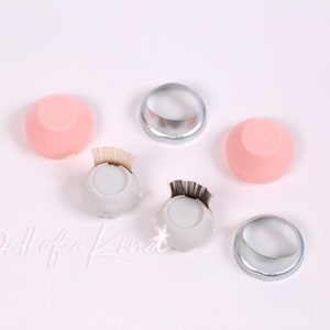 May include: A set of lash tools for applying false eyelashes. The set includes two pink silicone lash palettes, two white lash palettes with black lash strips, and two silver lash rings. The text "Doll of a Kind" is written on the white surface.