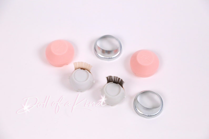 May include: A set of lash tools for applying false eyelashes. The set includes two pink silicone lash palettes, two white lash palettes with black lash strips, and two silver lash rings. The text "Doll of a Kind" is written on the white surface.