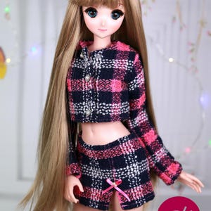Puede incluir: Una muñeca con un top corto de cuadros rosa y negro y una falda a juego. La muñeca tiene el pelo largo y castaño y los ojos azules.