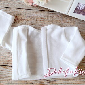 May include: White long-sleeved shirt with a snap closure down the back. The shirt is made of a soft, lightweight fabric. "Doll of a Kind" is printed on the bottom of the shirt.