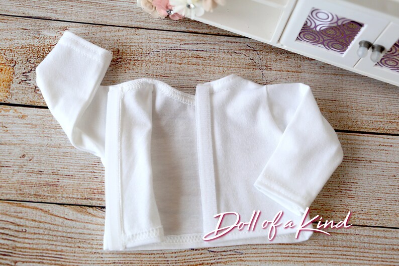 May include: White long-sleeved shirt with a snap closure down the back. The shirt is made of a soft, lightweight fabric. "Doll of a Kind" is printed on the bottom of the shirt.
