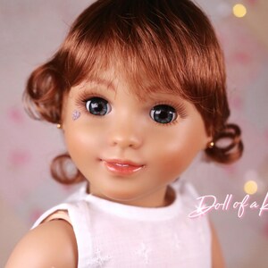 May include: A close-up of a doll's face with auburn hair styled in curls. The doll has large, blue eyes, long eyelashes, and a light complexion. A small, purple heart-shaped gem is on the doll's cheek. The text "Doll of a Kind" is in the bottom right corner.