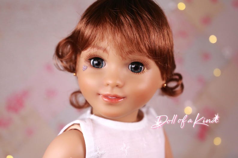 May include: A close-up of a doll's face with auburn hair styled in curls. The doll has large, blue eyes, long eyelashes, and a light complexion. A small, purple heart-shaped gem is on the doll's cheek. The text "Doll of a Kind" is in the bottom right corner.