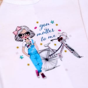 May include: White t-shirt with a cartoon girl wearing overalls and glasses, riding a bicycle. The text "you matter to me" is printed on the shirt.