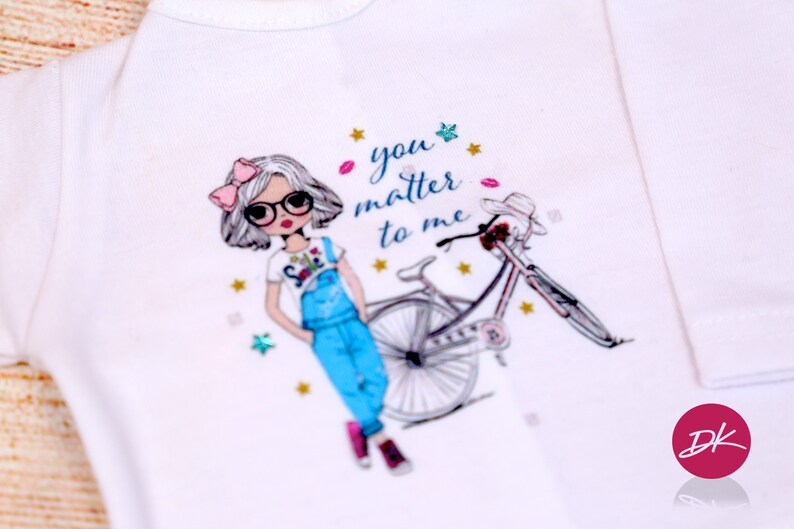 May include: White t-shirt with a cartoon girl wearing overalls and glasses, riding a bicycle. The text "you matter to me" is printed on the shirt.