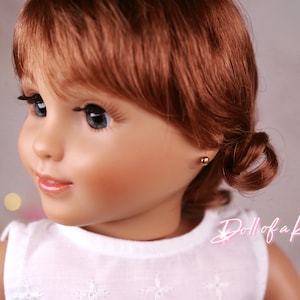 May include: A doll with auburn hair styled in a side bun. The doll has blue eyes, long eyelashes, and a fair complexion. It is wearing a white top and small gold stud earrings. The text "Doll of a Kind" is visible in the image.