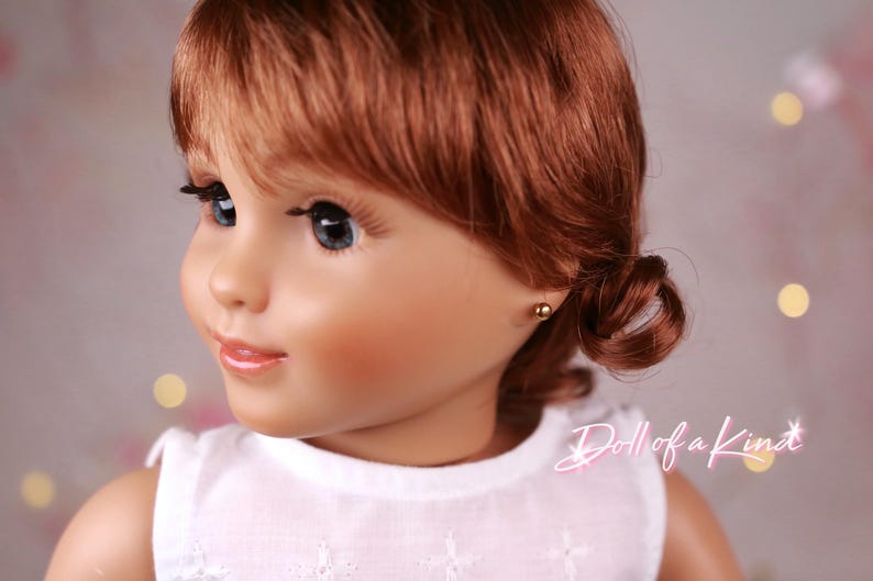 May include: A doll with auburn hair styled in a side bun. The doll has blue eyes, long eyelashes, and a fair complexion. It is wearing a white top and small gold stud earrings. The text "Doll of a Kind" is visible in the image.