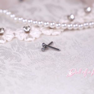 May include: A pair of small, silver-colored barbell earrings with a ball on each end, resting on a white, textured surface. A pearl necklace and lace trim with rhinestones are in the background. The text "Doll of a Kind" is in pink script.