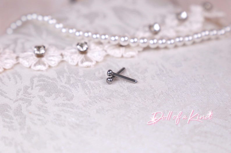 May include: A pair of small, silver-colored barbell earrings with a ball on each end, resting on a white, textured surface. A pearl necklace and lace trim with rhinestones are in the background. The text "Doll of a Kind" is in pink script.