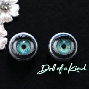May include: Two blue doll eyes with black eyelashes, set in silver metal casings. The eyes are set against a black background with the text "Doll of a Kind" in green.