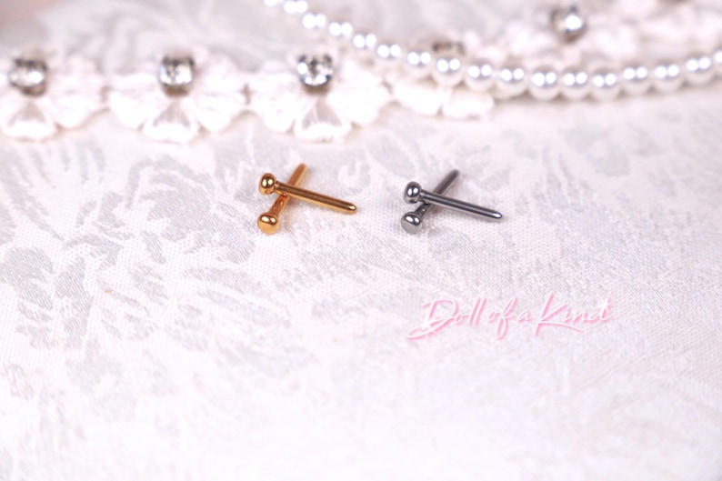 May include: Two metal body jewelry pieces. One is gold-toned, the other is silver-toned. Each has a rounded ball on one end and a straight post on the other. The jewelry is displayed on a white, textured surface with lace and pearl accents.