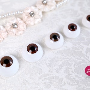 May include: Four brown glass doll eyes with black pupils, arranged in a row on a white surface. The eyes are different sizes.