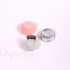 May include: Three eyelash application tools. A white tool with a black lash strip, a pink silicone tool, and a silver metal ring.