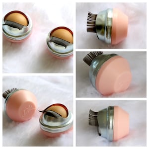 May include: Five pink and silver containers with a brush of black lashes sticking out of the top. The containers have a silver band around the middle and a pink lid. The containers are on a white surface.