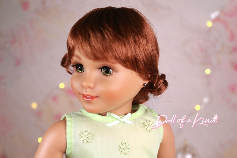 May include: A doll with auburn hair styled in pigtails and green eyes. The doll wears a light green dress with flower and bow details. The doll's face is detailed with makeup, and the background is softly blurred with bokeh lights.