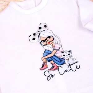 May include: White long-sleeve shirt with a graphic of a girl wearing glasses and a polka dot bow, sitting with a cat. The text "So Cute" is printed below the graphic.