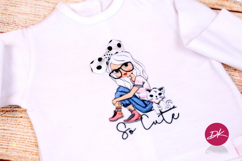 May include: White long-sleeve shirt with a graphic of a girl wearing glasses and a polka dot bow, sitting with a cat. The text "So Cute" is printed below the graphic.