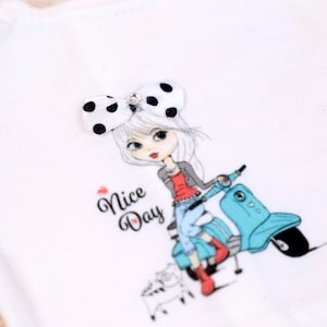 May include: White t-shirt with a graphic print of a girl riding a blue scooter. The girl has a black and white polka dot bow in her hair. The text "Nice Day" is printed on the shirt.