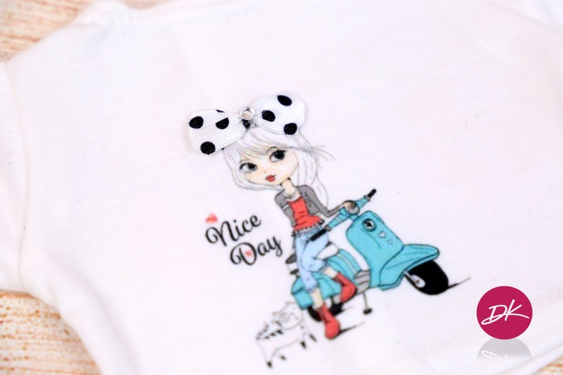 May include: White t-shirt with a graphic print of a girl riding a blue scooter. The girl has a black and white polka dot bow in her hair. The text "Nice Day" is printed on the shirt.