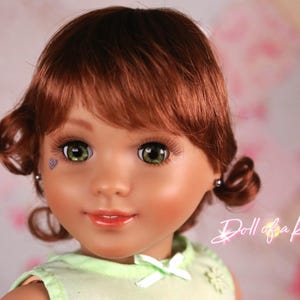 May include: A close-up of a doll with auburn hair styled in curls. The doll has green eyes, long eyelashes, and a small, decorative tattoo near one eye. The doll is wearing a light green top. The text "Doll of a Kind" is visible.