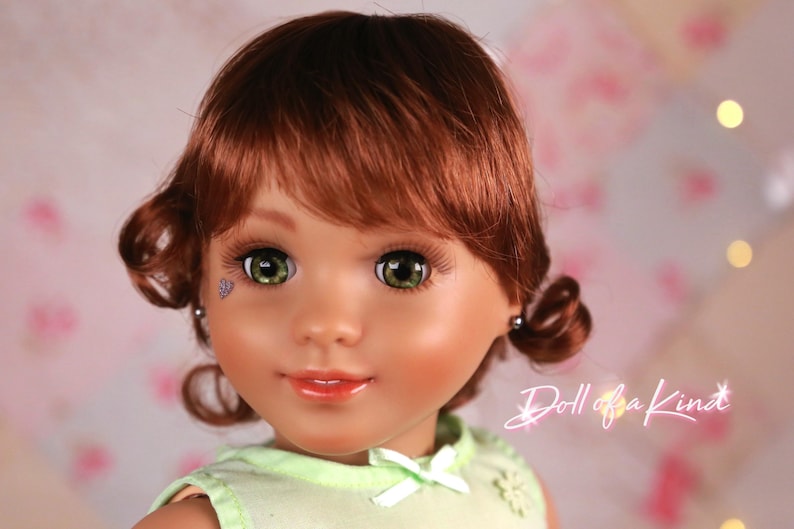 May include: A close-up of a doll with auburn hair styled in curls. The doll has green eyes, long eyelashes, and a small, decorative tattoo near one eye. The doll is wearing a light green top. The text "Doll of a Kind" is visible.