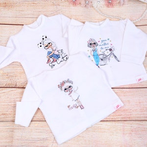 May include: Three white long-sleeved shirts with different graphic designs. The first shirt features a girl with a bow and a dog, with the text "So Cute". The second shirt features a girl riding a bicycle with the text "You matter to me". The third shirt features a girl with a flower crown and the text "The Girl".
