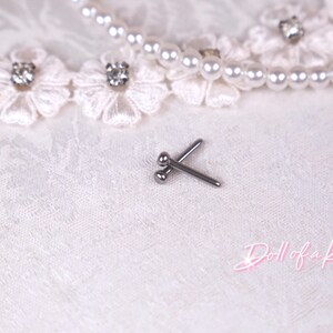 May include: A pair of small, silver-colored body jewelry studs. The studs are crossed and rest on a white, textured surface, with a pearl necklace and floral lace trim in the background. The text "Doll of a Kind" is in the lower right corner.