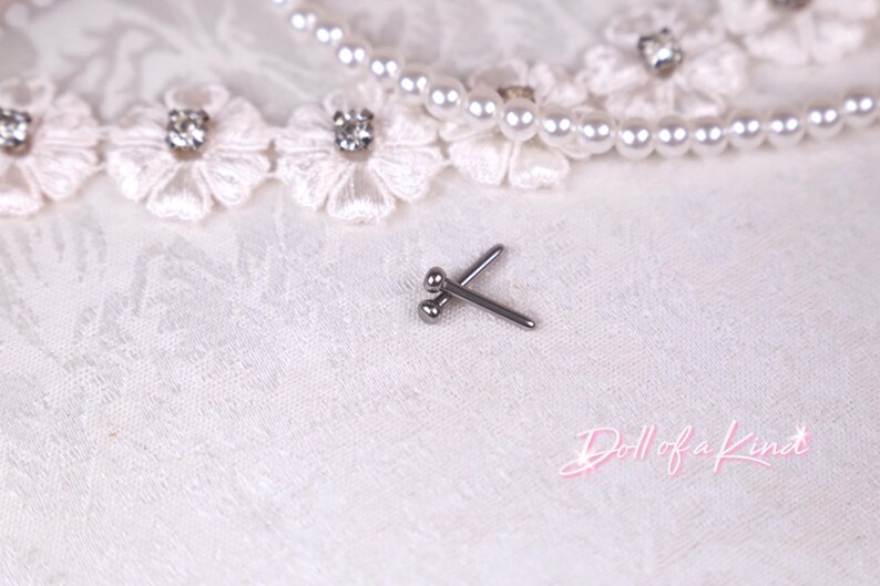 May include: A pair of small, silver-colored body jewelry studs. The studs are crossed and rest on a white, textured surface, with a pearl necklace and floral lace trim in the background. The text "Doll of a Kind" is in the lower right corner.