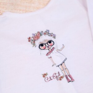 May include: White t-shirt with a graphic print of a girl wearing glasses and a flower crown. The text "Cut Girl" is printed below the graphic.