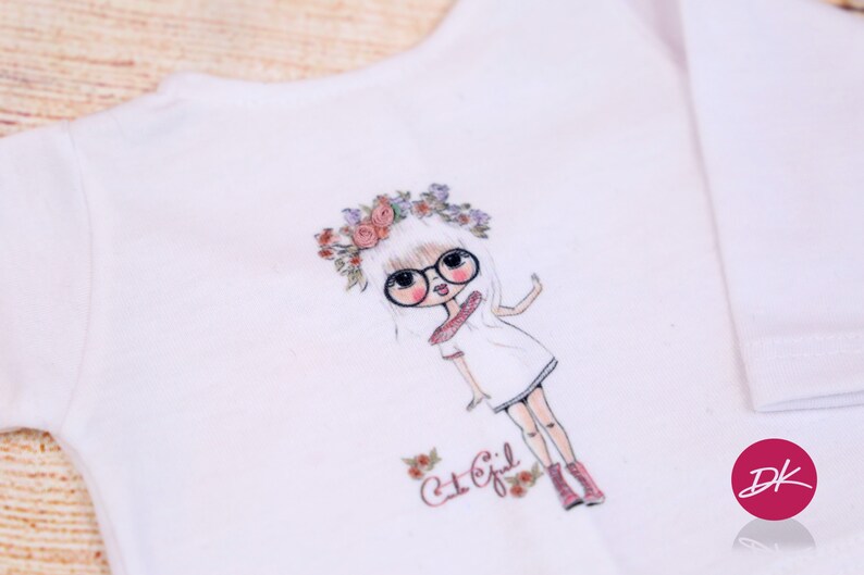 May include: White t-shirt with a graphic print of a girl wearing glasses and a flower crown. The text "Cut Girl" is printed below the graphic.