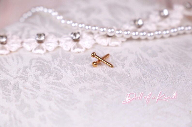 May include: A pair of gold-colored, barbell-style earrings with a ball on each end. The earrings are set against a white, textured fabric background, with a string of pearls and lace trim in the background.