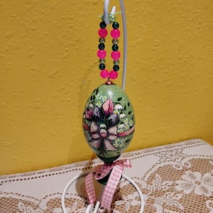 以下が含まれることがあります： 花柄とピンクのリボンで装飾されたイースターエッグのオーナメント。白い金属製のスタンドから吊り下げられており、ピンクとグリーンのビーズの連なりで飾られています。ピンクと白のチェック柄のリボンがベースに結ばれています。