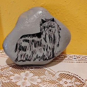 May include: A hand-painted rock featuring a black and white illustration of a dog. The dog has long, shaggy fur and is painted on a gray stone. The background is a solid yellow color.