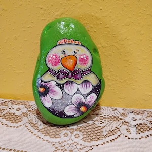 May include: Hand-painted rock art featuring a green egg-shaped stone with a chick design. The chick has a yellow crown, pink cheeks, and an orange beak. Purple flowers and a purple bow adorn the chick's eggshell.