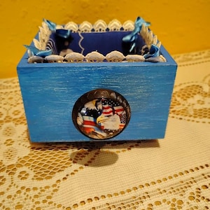 May include: A small, square, blue wooden box with a decorative oval emblem featuring an American flag and an eagle. The box is trimmed with white lace and blue ribbon. The box is sitting on a cream-colored lace tablecloth.