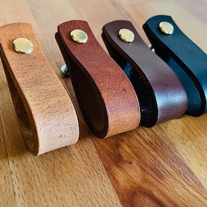 Puede incluir: Cinco tiradores de cajón de cuero en diferentes colores, incluyendo marrón, beige y negro. Cada tirador tiene un pomo redondo de latón.