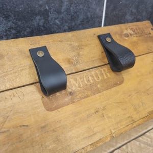 May include: Two black leather drawer pulls with brass rivets are attached to a weathered wooden surface. The pulls are looped and appear to be made of a smooth leather. The wooden surface has the word "MOUR" stamped on it.