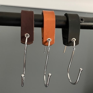 May include: Three leather strap hooks in brown, tan, and black, each with a silver-colored metal S-hook. The hooks are hanging from a black metal bar, offering a simple and stylish storage solution.