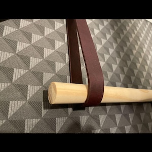May include: A wooden dowel with a burgundy leather strap, designed for hanging items. The dowel is light-coloured, and the strap is looped around it. The background is a grey and white geometric pattern.
