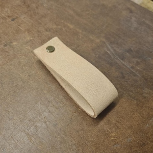 May include: A light tan leather pull tab with a brass rivet. The loop is curved and appears to be made of suede or a similar textured material. The leather is a neutral colour, suitable for various applications.
