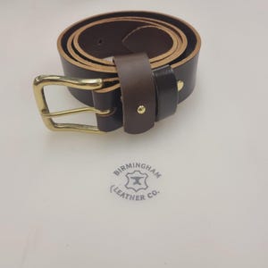 May include: A rolled-up brown leather belt with a gold-colored buckle. The belt is dark brown and has a classic design. The image also shows the logo of Birmingham Leather Co.